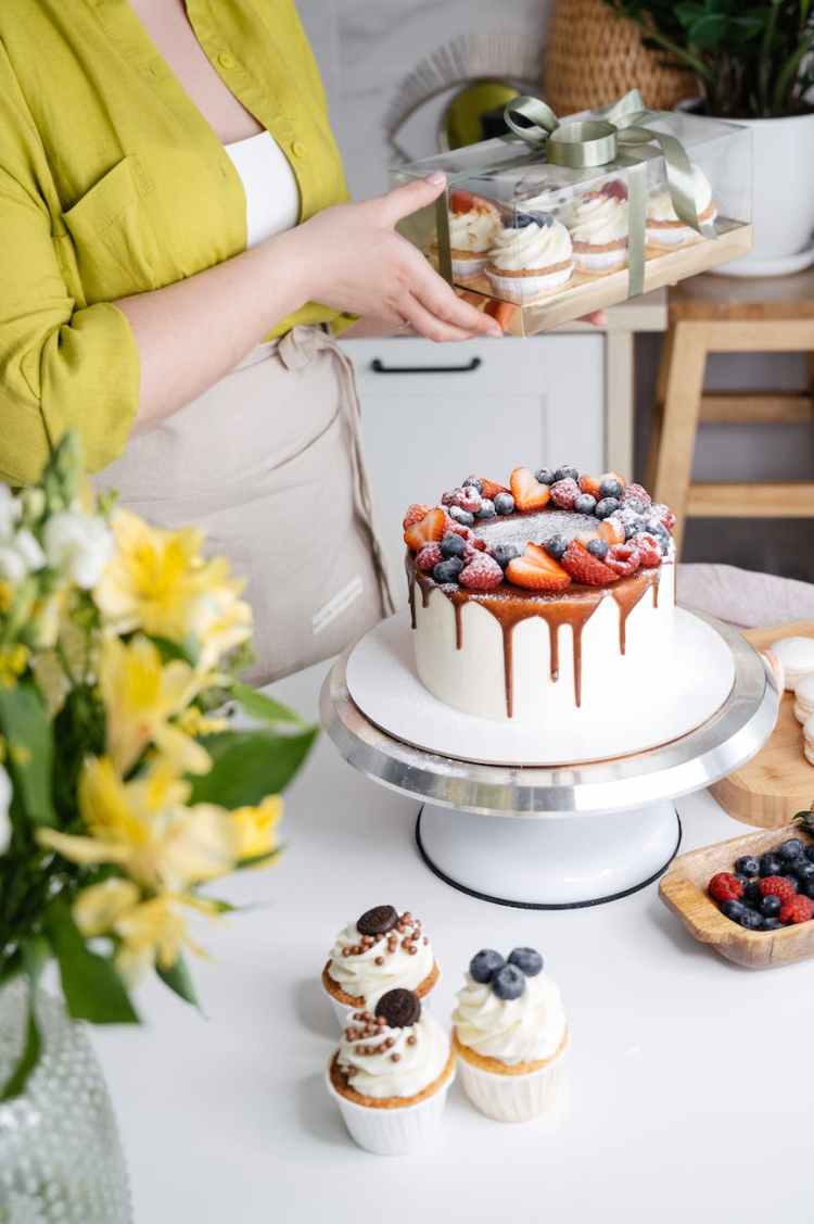 woman with decorated cake on stand and cupcakes in box
